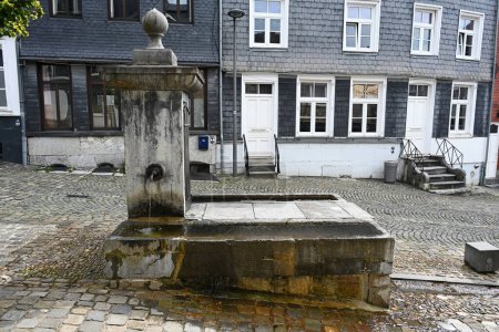 Antigua fuente de grifo de agua pública en una plaza frente a fachadas cubiertas de azulejos de pizarra de auténticos hogares belgas. Fuente en el centro histórico de Stavelot, Bélgica