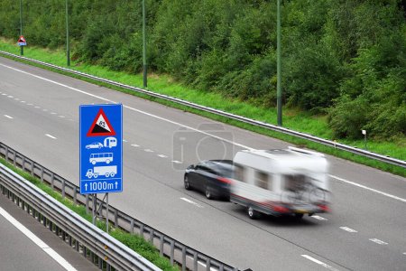 Auto mit Reiseanhänger passiert Verkehrsinformationsschild auf der Autobahn E42, der Route durch die belgischen Ardennen bei Malmedy