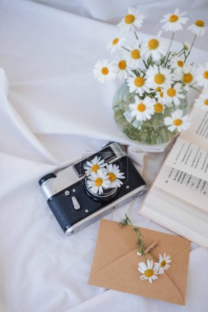 Photo for Chamomile flowers in vase, vintage camera and book on white fabric. - Royalty Free Image