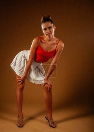 Jolie jeune femme en posture penchée vers l'avant. Elle porte un haut rouge, une jupe blanche et des talons hauts