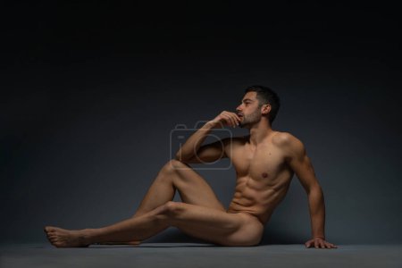 Mode-Nacktfoto eines männlichen Modells mit verführerischer Figur, das isoliert auf dem Boden in einem Studio sitzt