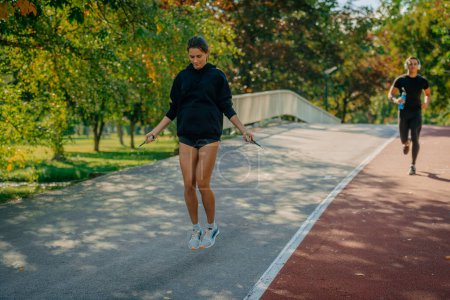 A male sportsperson is running a marathon around the park while listening music on his headphones. His girlfriend is jumping rope