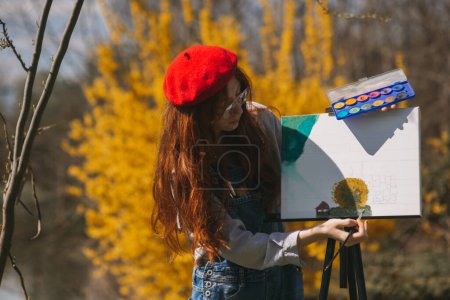 Beautiful ginger girl with red beanie and glasses painting on a canvas while standing at the park