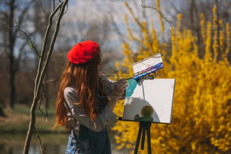 Ginger girl with red hat painting on an easel with her color palette while standing in front of the easel