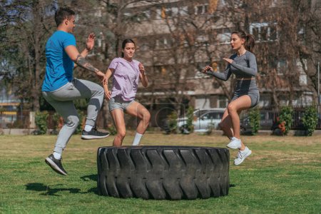 Beautiful, young sports people jumping on big truck tire in the park. They are exercisnig their legs