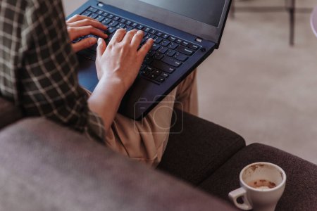 Employée travaillant sur un ordinateur portable alors qu'elle était assise sur un canapé gris avec une tasse de café à côté d'elle. Photo rapprochée