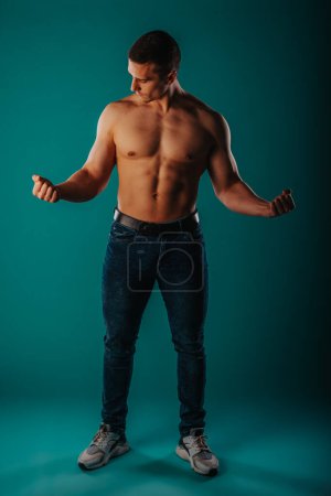Hemdloser Typ, der seinen fitten Körper zeigt, Bizeps, Brust, Bauch. Starker Mann blickt auf seinen Bizeps, während er vor türkisfarbenem Hintergrund im Studio posiert