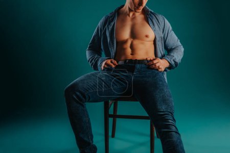 Hombre deportivo sentado en una silla con camisa desabotonada posando sobre un fondo turquesa en el estudio