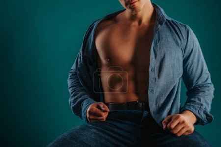 Male sports person showing his fit torso posing while sitting on a chair with unbuttoned shirt. Partial photo.