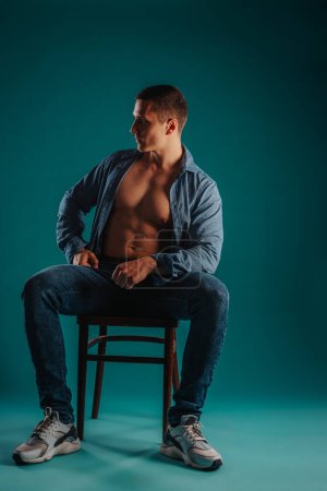 Fit shirtless guy with unbuttoned shirt sitting on a chair posing in studio on a turquoise background. He is looking aside.