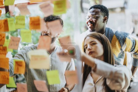 Creative professionals work on a project, utilizing a glass board and sticky notes. Successful teamwork and communication lead to a modern, persistent corporation.