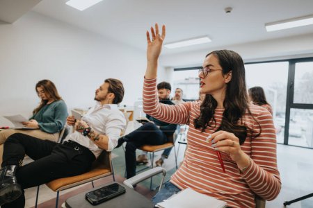 Jeune femme focalisée avec des lunettes levant la main dans un cadre de classe avec des étudiants attentifs et un environnement éducatif moderne.