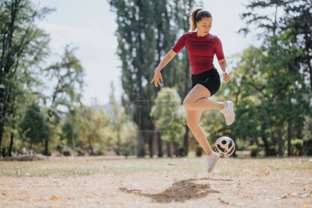 Attractive athlete enjoys training outdoors with a soccer ball, performing football freestyling tricks on a sunny day in a park. The energetic girl embraces a sporty and healthy lifestyle in nature.