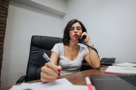 Focused businesswoman multitasking with phone and taking notes at her desk in a modern office setting.