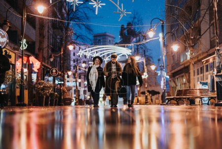 A group of young adults strolls down a wet city street adorned with festive lights, reflecting the urban vibe and companionship.