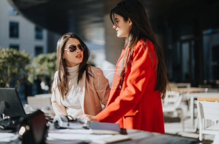 Fesselndes Bild, das zwei junge professionelle Frauen tief im Gespräch in einem sonnigen Outdoor-Café zeigt, wobei eine Frau selbstbewusst gestikuliert.