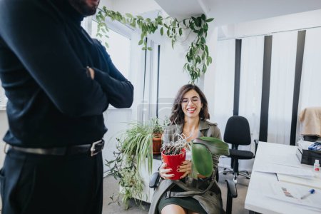 A happy woman holding a potted plant laughs while interacting with a male colleague in a well-lit, contemporary office space.