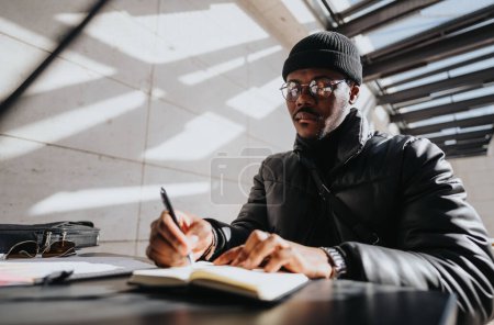 An African American man with glasses, wearing a beanie and a winter jacket, pensively jotting down notes in a journal at a modern workspace.