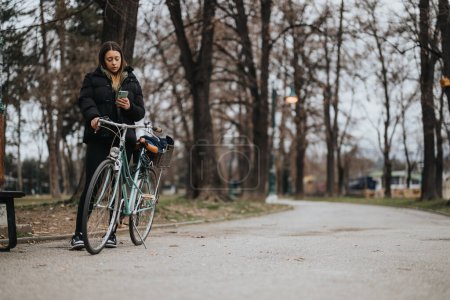 A focused young adult checks her phone after a bike ride. Fall atmosphere in a peaceful park, with trees and a path in the background.