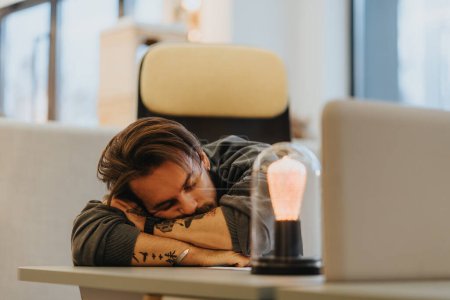 Overworked young adult napping at his desk with a laptop in a contemporary office. The image captures the concept of workplace fatigue and the need for work-life balance.