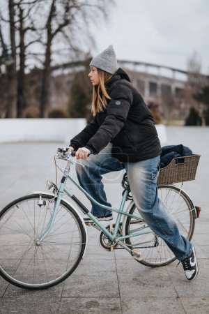 Focused young adult pedaling a retro-style bike with a basket in a city park on a chilly day, showcasing an active lifestyle.