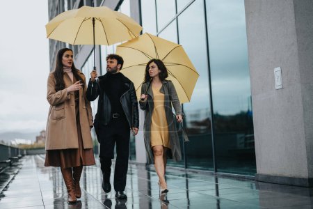 Eine Gruppe von drei jungen erwachsenen Freunden spaziert und unterhält sich unter zwei gelben Regenschirmen auf einem nassen, reflektierenden Stadtweg..