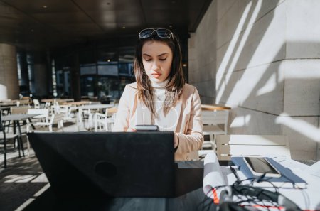 Junge Geschäftsfrau bei der Arbeit an ihrem Laptop in einem Café im Freien, gebadet im Sonnenlicht, mit einem Smartphone in der Hand.