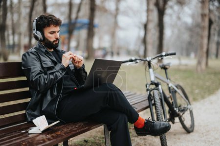 Focused businessman in stylish outfit remotely working on laptop with headphones, sitting on park bench near his bicycle.