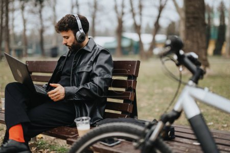 Stylish businessman enjoys remote work on his laptop while sitting on a bench in a city park, with his bicycle beside him.