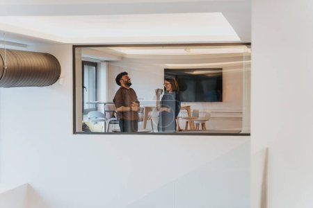 Un hombre y una mujer son vistos en un momento franco, charlando y riendo en un ambiente de oficina contemporáneo a través de una ventana.