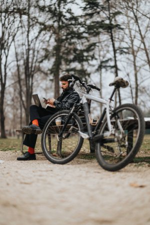 Ein modischer männlicher Unternehmer erledigt geschäftliche Aufgaben auf seinem Laptop in einem ruhigen Park, das Fahrrad an seiner Seite.