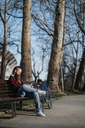Homme d'affaires barbu en tenue décontractée utilisant une tablette à l'extérieur, avec un vélo et un casque par une journée ensoleillée dans une zone de parc urbain.