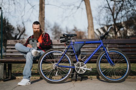 A bearded businessman in casual wear working on a tablet, with his bicycle beside him, in an urban park setting.