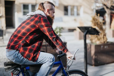 Hipster-Geschäftsmann auf einem blauen Fahrrad in einem Stadtgebiet, der mit seinem Smartphone ferngesteuert und mit urbanem Lifestyleflair zur Arbeit fährt.