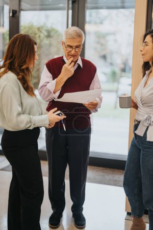 Experienced senior executive ponders over paperwork with attentive female colleagues in a well-lit modern office setting, collaboration in action.