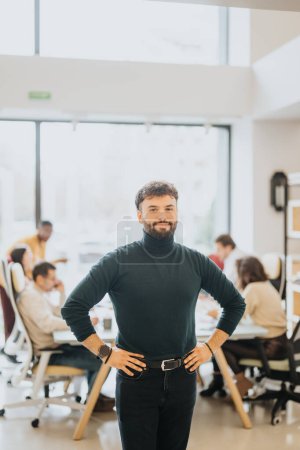 Young, assured male professional in a turtleneck stands hands-on-hips in an office with colleagues working in the background.