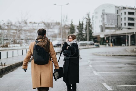 Rückansicht eines trendigen jungen Mannes und einer jungen Frau in Wintermänteln, die auf einer schneebedeckten Straße mit urbanem Hintergrund spazieren.