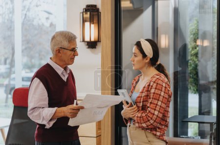 An experienced senior man in spectacles engaged in a discussion with a younger female colleague holding a document and a digital tablet
