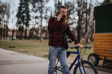 Entrepreneur masculin élégant utilisant le téléphone intelligent tout en se tenant avec son vélo dans un parc urbain, mettant en valeur le travail à distance moderne.