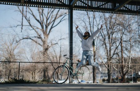 Joyful young woman jumping in the air beside bicycle in urban park.