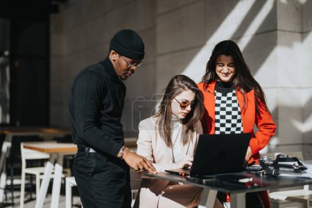 Drei junge, multiethnische Menschen kollaborieren mit einem Laptop an einem sonnigen Café-Tisch im Freien und zeigen Teamwork und Kreativität.