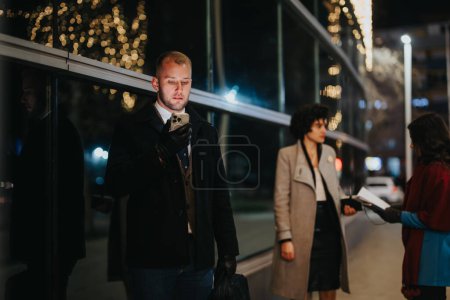 Commuters waiting at a bus stop on a bustling city night. Focused man with smart phone in foreground. Blurred lights backdrop.