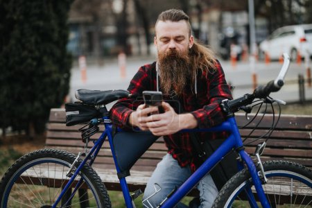 Stilvoller bärtiger Mann mit langem Bart, der auf einer öffentlichen Bank sitzt und sein Handy kontrolliert, neben ihm ein blaues Fahrrad..