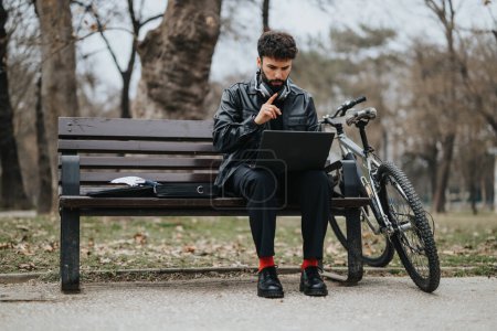 Ein fokussierter männlicher Unternehmer, der in einem Park mit einem Fahrrad neben sich an einem Laptop arbeitet und eine Mischung aus Professionalität und aktivem Lebensstil verkörpert.
