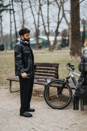 Un hombre de negocios con estilo en un abrigo negro que trabaja al aire libre con una bicicleta, ilustrando el trabajo remoto en un entorno de parque urbano natural.