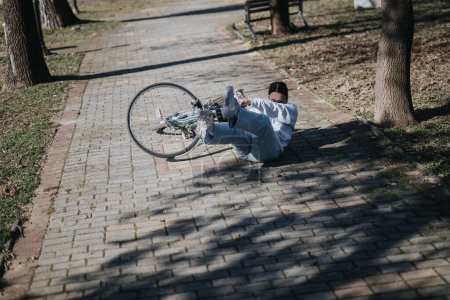 Young adult in casual clothing experiencing a bicycle accident on a paved park pathway.