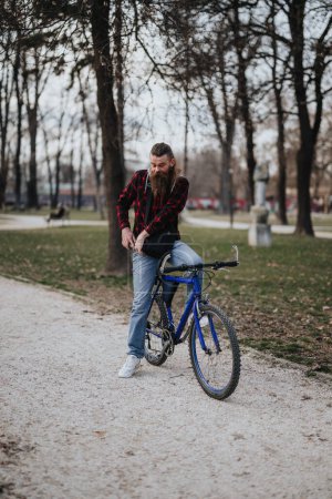 A hipster man with a long beard pauses on his bike in a tranquil park setting to use his phone.