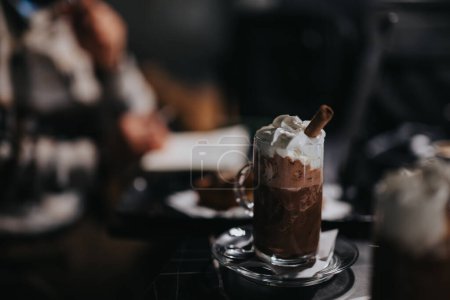 Cozy coffee shop vibe with a focus on a delicious hot chocolate topped with whipped cream and cinnamon stick.