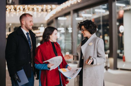 Outdoor evening discussion as a smiling real estate agent shares property information with an interested couple outside a business venue adorned with festive lights.