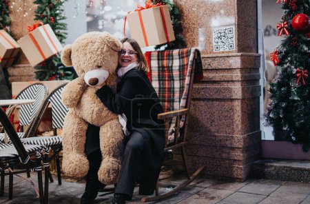 Smiling woman holding a big teddy bear with festive Christmas decorations and gifts around on a cozy cafe terrace.